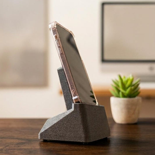 A gray textured triangular phone stand holding a smartphone, placed on a dark wooden table with a small potted succulent and a blurred office background.